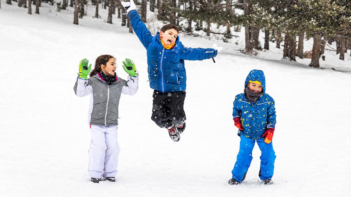 Children in snow pants and jackets jumping in the snow in front of a forest