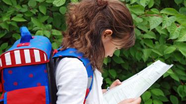An elementary school student with medium length brown hair examines her report card while walking along with her backpack on in front of a green, leafy bush.