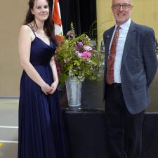 Young woman in long sleeveless long navy gown stands beside man in grey suit in front of a vase of flowers